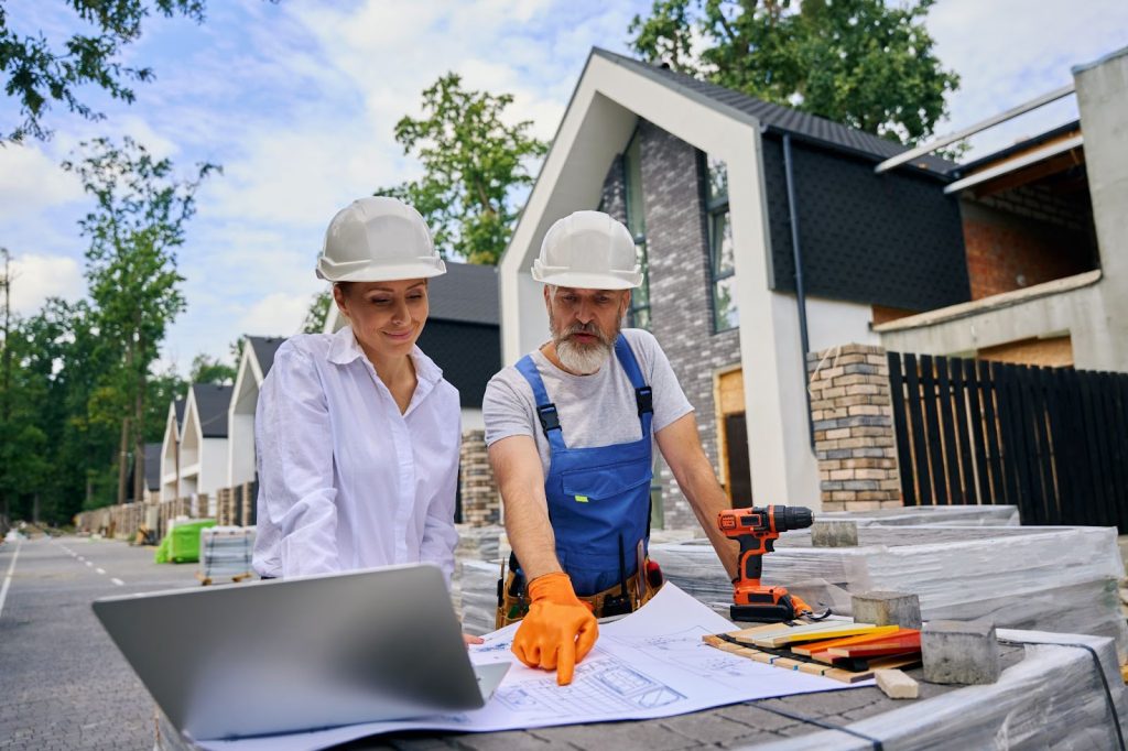 Builder pointing at architectural drawings while discussing outdoors with a pleased construction supervisor.