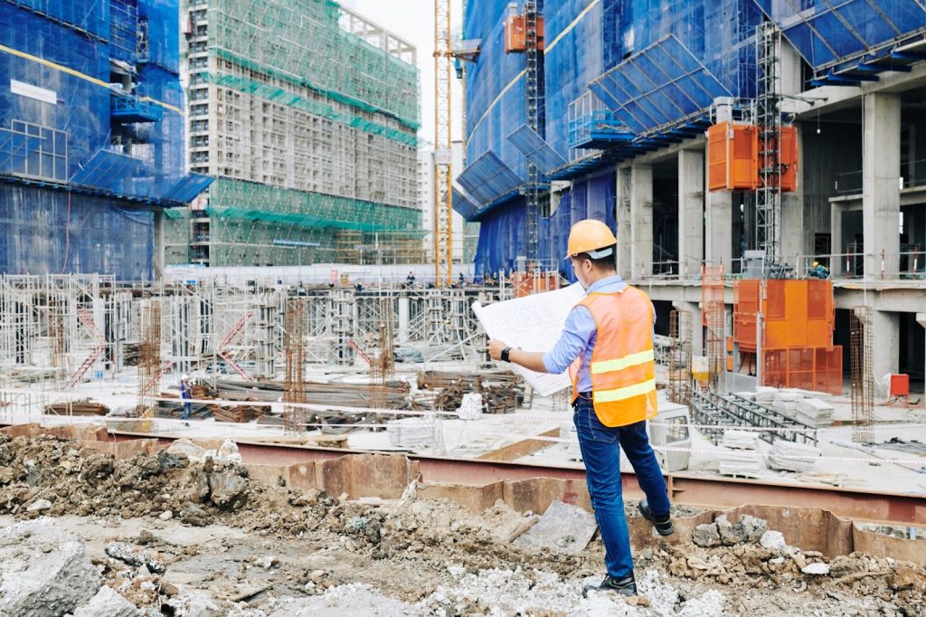 Rear view of the head engineer in an orange vest examining the building blueprint when standing at the construction site.