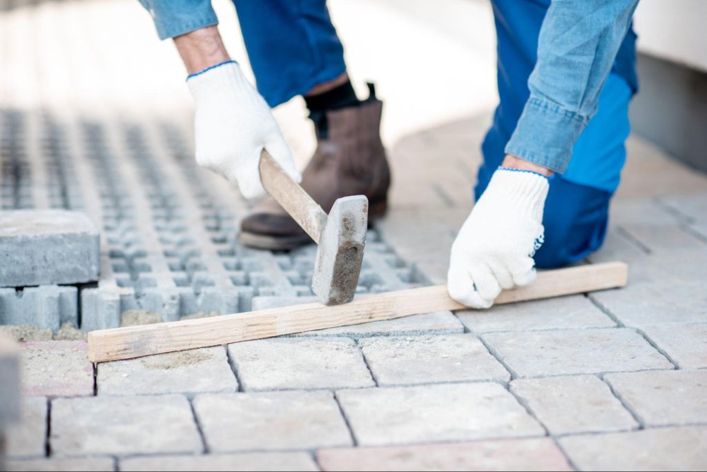 A builder's hand in white gloves lays paving tiles on a sidewalk undergoing repairs; his face is not in view.
