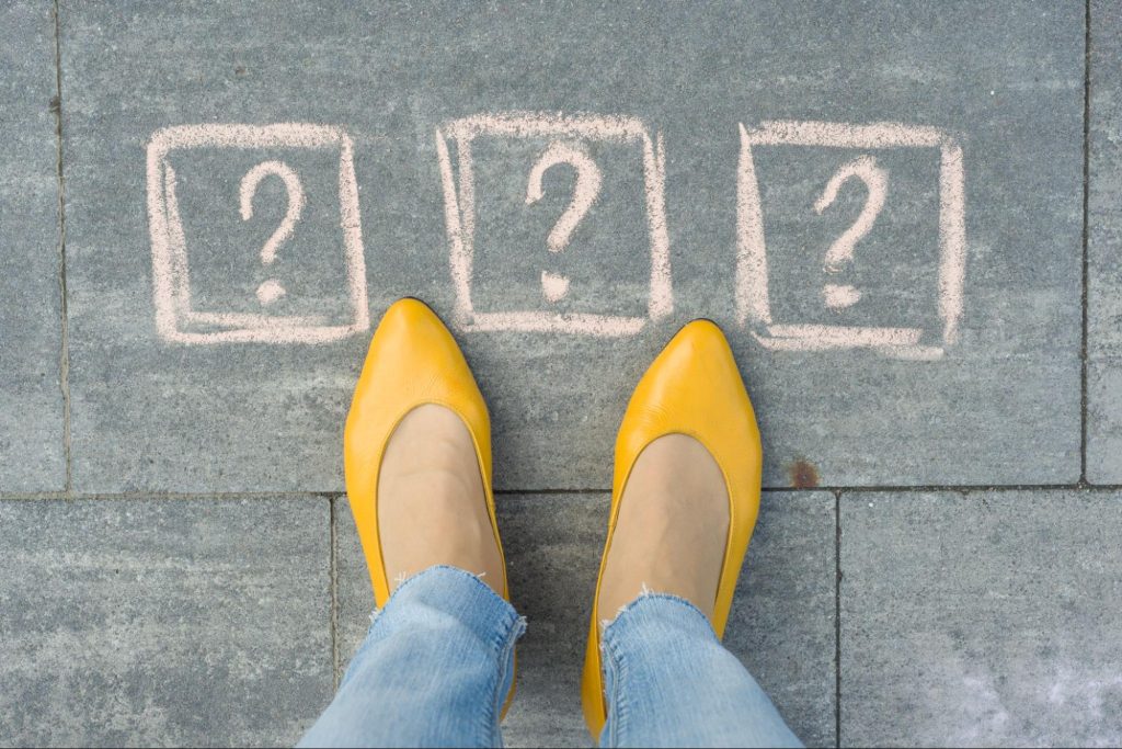 An image of a woman's feet in mustard pumps standing on a grey sidewalk with three question marks painted before her, illustrating the questions to ask an asphalt paving contractor.
