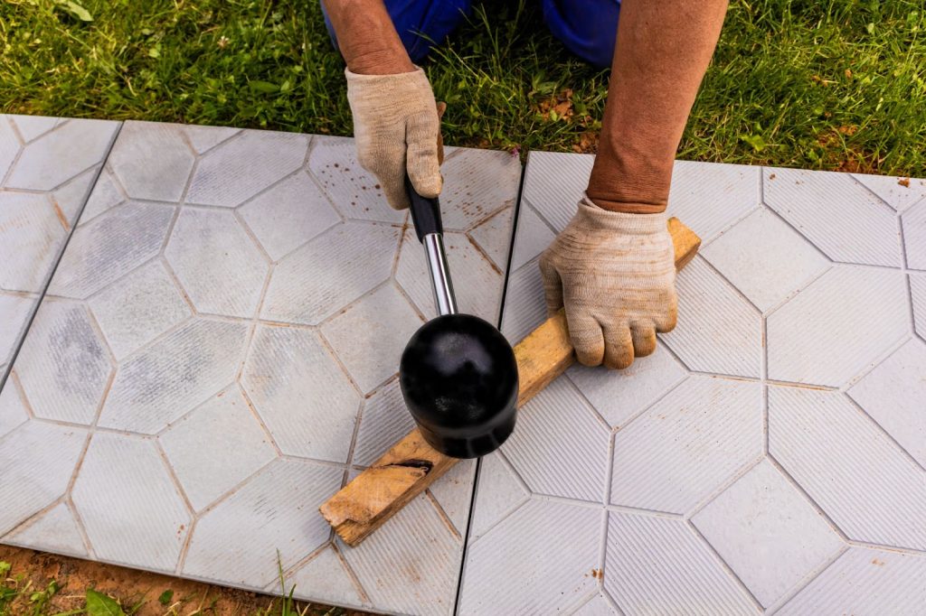 The hands of a professional worker hit the slab with a special rubber hammer while laying a garden path made of stone tiles.