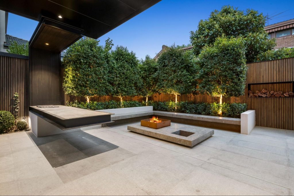 A modern backyard concrete patio featuring sleek concrete benches and a bar table, surrounded by trees and against a backdrop of walls with wooden designs, creating a contemporary outdoor setting.