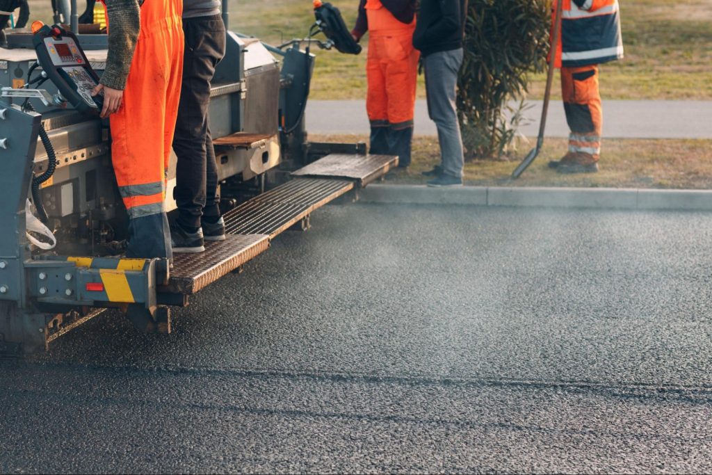 A paver machine laying fresh asphalt on a city street, with workers guiding and smoothing the surface.