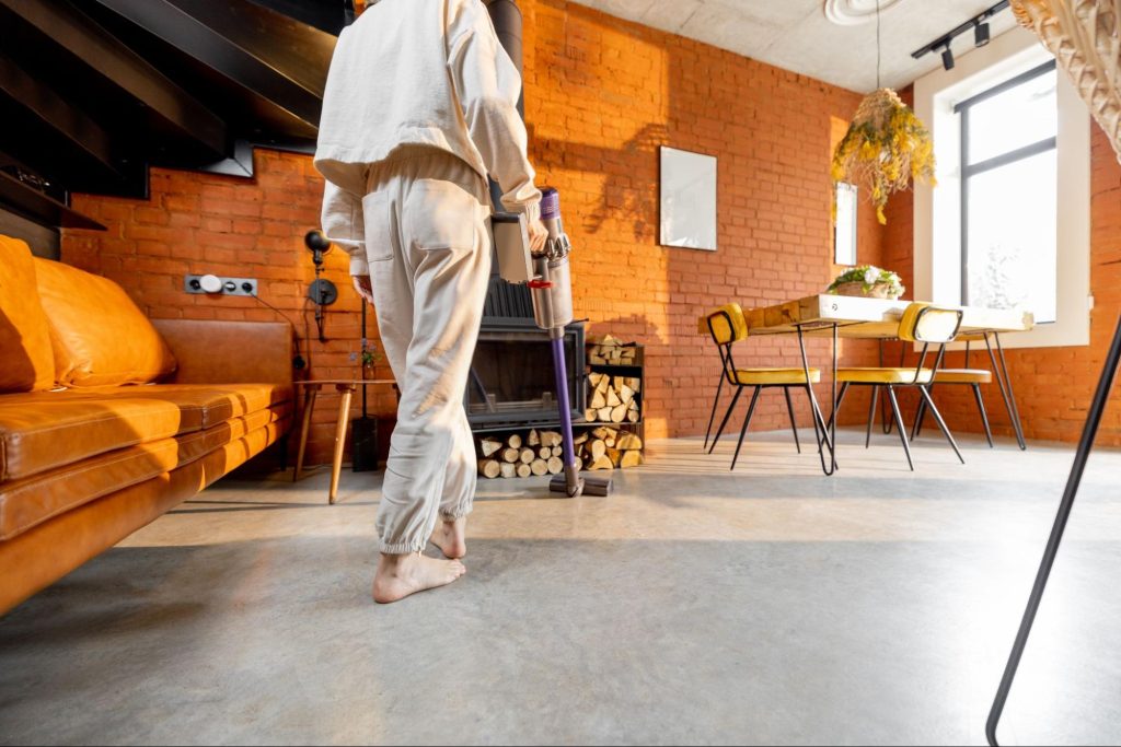 A woman vacuums a concrete floor in a cozy brick-walled room with an orange sofa, dark wooden stairs, a chimney with wood logs, and a wooden table with chairs.
