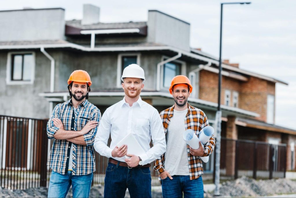 Three smiling residential concrete contractors in hard hats pose at the camera on a building site street, ready for construction work.